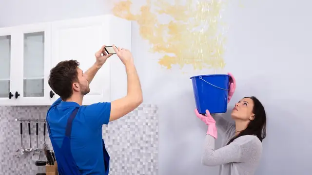 Homeowner photographing a water-stained wall while another person holds a bucket under the leak
